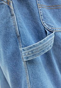 Blå denimstof med en synlig baglomme og en strop af stof. Syninger i en kontrasterende farve fremhæver detaljerne og strukturen.