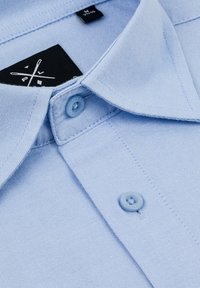 Jasnoniebieska koszula z kołnierzykiem wykonana z teksturowanej tkaniny, z dwoma jasnoniebieskimi guzikami i czarną metką wszytą wewnątrz kołnierza.