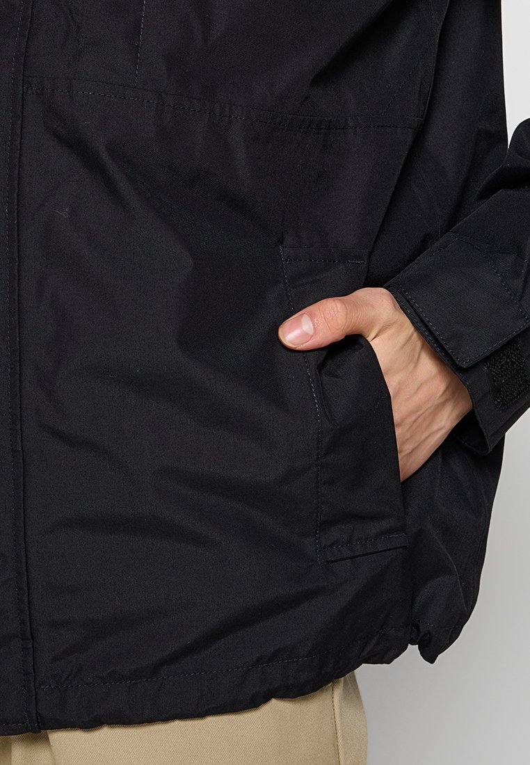 Chaqueta negra con una textura suave, que cuenta con bolsillos laterales y puños de velcro. La tela es ligera y resistente al agua.
