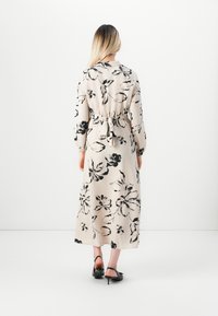 Femme aux longs cheveux blonds portant une robe maxi beige avec des imprimés floraux noirs nouée à la taille, se tenant en talons noirs sur fond blanc.