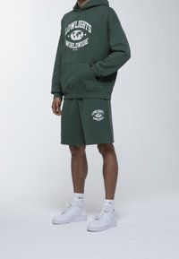 Sudadera verde oscuro con gráfico blanco, pantalones cortos a juego y zapatillas blancas. Tela de mezcla de algodón, corte relajado, bolsillo frontal, detalle de logo en los pantalones cortos.