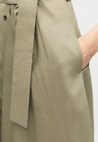 Χέρι στην τσέπη ενός φορέματος σε ελιά πράσινο με ψιλή ρίγα, ζώνη από το ίδιο ύφασμα δεμένη στη μέση και μαύρες λεπτομέρειες με κουμπιά.