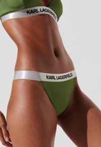 Žalias bikini apatinis trikotažas su sidabriniu elastingu dirželiu, ant kurio juodu šriftu užrašyta "KARL LAGERFELD". Lygi tekstūra, tanga dizainas.