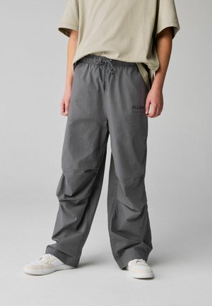 Person wearing loose gray drawstring pants with black text on thigh, beige oversized T-shirt, and white sneakers standing against a plain background.