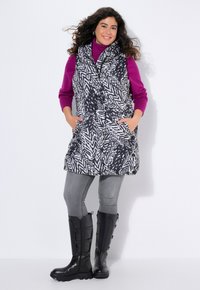 Zwart-wit gestreept vest met hoge kraag, twee zijzakken, gelaagd over een paarse coltrui, gecombineerd met grijze skinny jeans en zwarte laarzen.