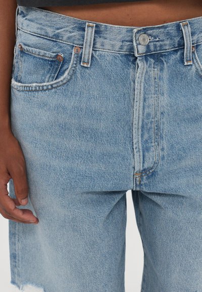 Jeans en denim bleu clair à taille haute, cinq poches et coutures visibles. Présentent un ourlet effiloché et des accessoires en ton cuivré.