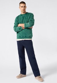 Sudadera de algodón verde con puños y cuello de canalé, superpuesta a una camisa beige. Combinada con pantalones azul marino y zapatillas beige.