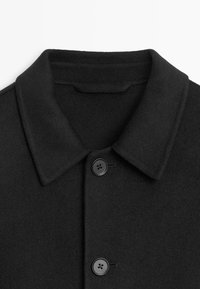 Schwarzer Wollmantel mit einem Kragen, ausgestattet mit zwei schwarzen Knöpfen vorne und einem glatten, strukturierten Stoff.