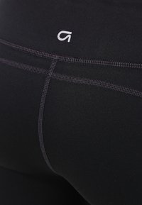 Svarta sportleggings med en slät textur, med en diskret logotyp och dekorativa sömmar längs sömmarna för detaljer.