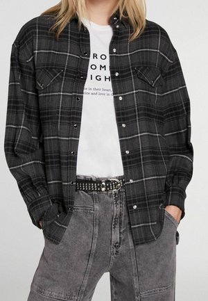 Kobieta ubrana w czarno-szarą koszulę w kratę na guziki, nałożoną na białą koszulkę z nadrukiem, szare dżinsy z wysokim stanem i czarny pasek z nitami.