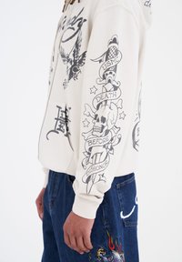 Sudadera crema con diseños gráficos en negro que incluyen motivos de águila y calavera. Presenta un cierre frontal y bolsillos laterales. Se lleva con pantalones de mezclilla azules.