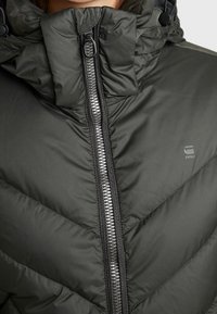 Mörkgrön, vadderad jacka med hög krage, dragkedjestängning och sicksackmönstrad sömnad. Har en logga på bröstet och ett strukturerat tyg.