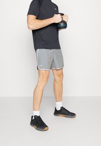Man i svart skjorta och grå shorts som håller en kettlebell, står på ett grått golv med vit bakgrund.
