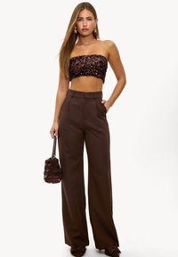 Haut court marron à sequins et pantalon marron taille haute avec plis. Accessoires comprennent un petit sac texturé. Mannequin debout, une main sur la hanche.