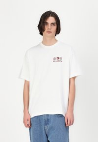 Balta medvilnė T-shirt su laisvu kirpimu, puošta raudonais grafika, įskaitant medžius ir žodį "BILLABONG" kairėje krūtinės pusėje.