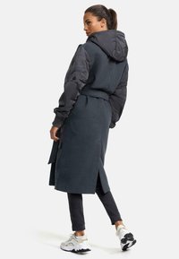 Long dark gray coat with a hood, featuring padded sleeves, a tie waist, and side slits. Paired with dark pants and gray sneakers.