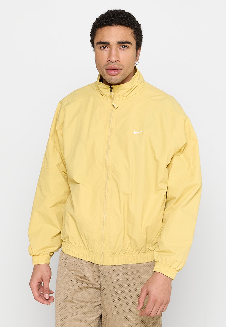 Lys gul nylon zip-jakke med høj krave, elastisk talje og manchetter; har et lille hvidt Nike-logo på venstre bryst.