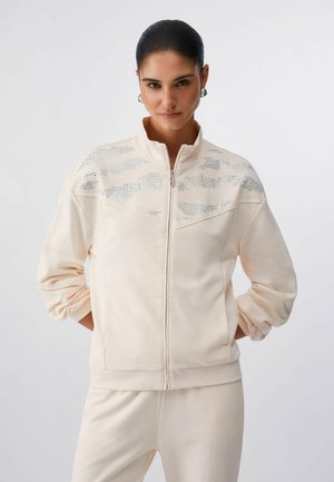 Femme aux cheveux foncés coiffés en chignon élégant, portant une veste crème à fermeture éclair ornée de motifs en clous argentés et un pantalon crème assorti, debout devant un fond blanc uni.