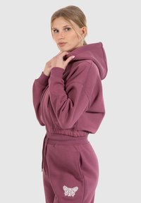 Completo con felpa color bordeaux e pantaloni abbinati. Tessuto morbido, design accorciato, tasca a marsupio e dettaglio grafico di farfalla sui pantaloni.