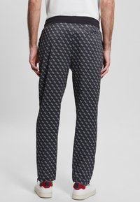 Muž v černých kalhotách se vzorem a bílými logy, v bílých teniskách se červenými a modrými detaily, stojící na jednobarevném šedém pozadí.