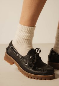 Άτομο που φοράει λευκές ριγωτές κάλτσες και μαύρα, υφασμένα δερμάτινα παπούτσια με κορδόνια και παχιές καφέ λαστιχένιες σόλες.