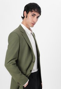 Blazer verde oliva con acabado texturizado, diseño de un solo botón y dos botones. Llevado sobre una camisa blanca impecable y pantalones negros.