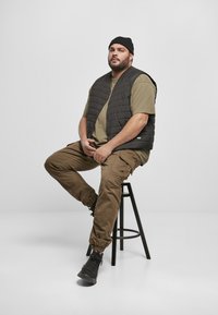 Svart polstret vest over en olivengrønn skjorte, kombinert med brune cargo-bukser og svarte joggesko, sittende på en svart krakk mot en hvit bakgrunn.