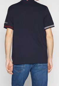 Námořnicky modré polo tričko s krátkým rukávem a proužkovanými akcenty na rukávech, s klasickým límečkem a malým logem na levé paži.