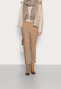 Beżowa kurtka puchowa, jasnobeżowy sweter, jasnobrązowe spodnie z rozcięciami po bokach, brązowe botki za kostkę oraz jasny szalik z frędzlami.