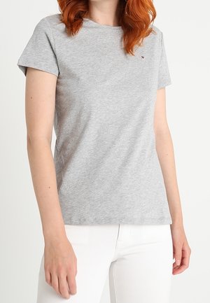 Femme aux cheveux roux portant un t-shirt uni gris clair à manches courtes et un pantalon blanc, debout sur un fond blanc.