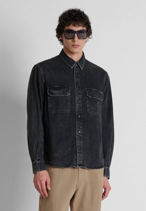 Junger Mann mit großen schwarzen Sonnenbrillen, schwarzem ausgewaschenem Denim-Hemd mit Brusttaschen und beigen Hosen vor schlichtem grauem Hintergrund.