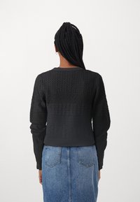 Černý pletený svetr se strukturovanými vzory a přiléhavým pasem, doplněný modrou džínovou sukní.