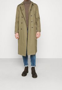 Olivgrüner Trenchcoat mit doppelter Knopfleiste, Knopfdetails und Schulterklappen, kombiniert mit einem braunen Rollkragenpullover, Jeans und braunen Stiefeln.