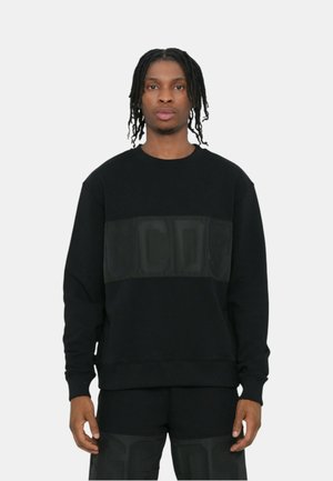 Giovane uomo con capelli intrecciati indossa felpa nera e pantaloni con grandi lettere in rilievo sul davanti, su sfondo uniforme.