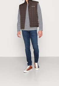 Brun vadderad väst med ståkrage över en navy- och vitrandig långärmad skjorta, ihop med mörkblå jeans och flerfärgade sneakers.