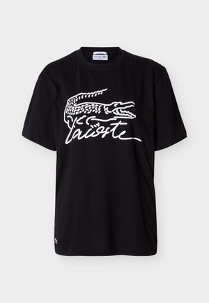 Tricou negru cu mânecă scurtă, cu un logo mare alb Lacoste care prezintă un crocodil stilizat și numele brandului scris în stil cursiv pe partea din față.