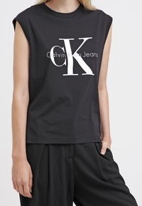 Vrouw in een mouwloze zwarte Calvin Klein Jeans top met groot wit "cK" logo, gecombineerd met losse zwarte broeken.
