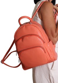 Zaino in pelle corallo dalla forma arrotondata, con due tasche frontali con zip e spallacci regolabili. Tessuto liscio con dettagli di cucitura discreti.