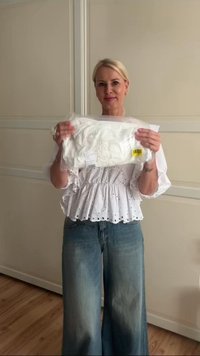 White, eyelet peplum top held in front, accompanied by medium-wash jeans. The background features light wood flooring and neutral walls.