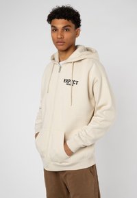 Beige zip-up hoodie with drawstring hood, front pockets, and printed text "EXPECT NOTHING" in black on left side. Soft material and relaxed fit.