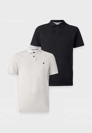 Uomo in piedi con le mani in tasca che indossa una polo nera a maniche corte, pantaloni cargo neri e scarpe nere su sfondo bianco uniforme.