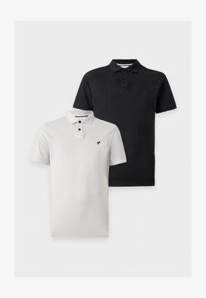 Uomo in piedi con le mani in tasca che indossa una polo nera a maniche corte, pantaloni cargo neri e scarpe nere su sfondo bianco uniforme.