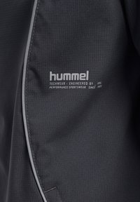 Svart techwear-jacka med ett texturerat tyg som har en subtil rutmönster. Inkluderar synlig branding och grå detaljer längs sömmarna.