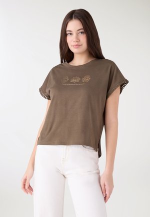 Giovane donna che indossa una maglietta marrone a maniche corte con frange e pantaloni bianchi, con tre simboli dorati e del testo sul petto.