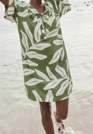 Personne portant une robe verte avec de grands motifs de feuilles blanches, debout dans une eau peu profonde, les bras levés au-dessus de la tête.