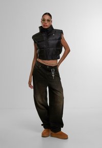 Black cropped puffer vest with high collar, paired with dark baggy jeans featuring a keychain detail. Model wearing brown platform shoes.