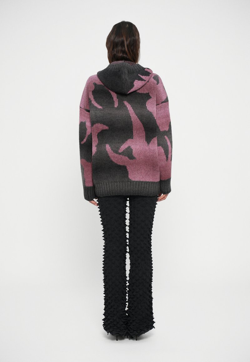 Felpa over-size grigia e rosa lavorata a maglia con un motivo audace, abbinata a pantaloni neri svasati testurizzati con dettagli in stile 3D.