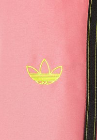 Rosa tyg har en broderad gul logga med tre bladdelningar. Svarta vertikala ränder med gul sömnad syns på sidan.