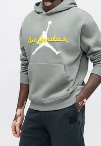 Personne portant un sweat à capuche Air Jordan gris avec écriture jaune et logo blanc, associé à un pantalon foncé, mains partiellement dans les poches.