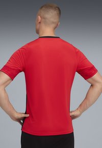 Rød atletisk trøje med korte ærmer, strukturerede skulderdetaljer og sort kant ved halsen. Minimalistisk design, tætsiddende form.